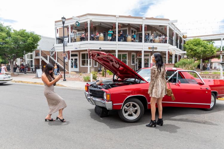 A young woman takes a picture of another young woman in front of an old car